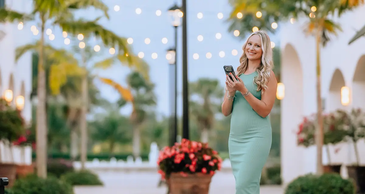 Elena smiling with her phone beneath string lights and palm trees